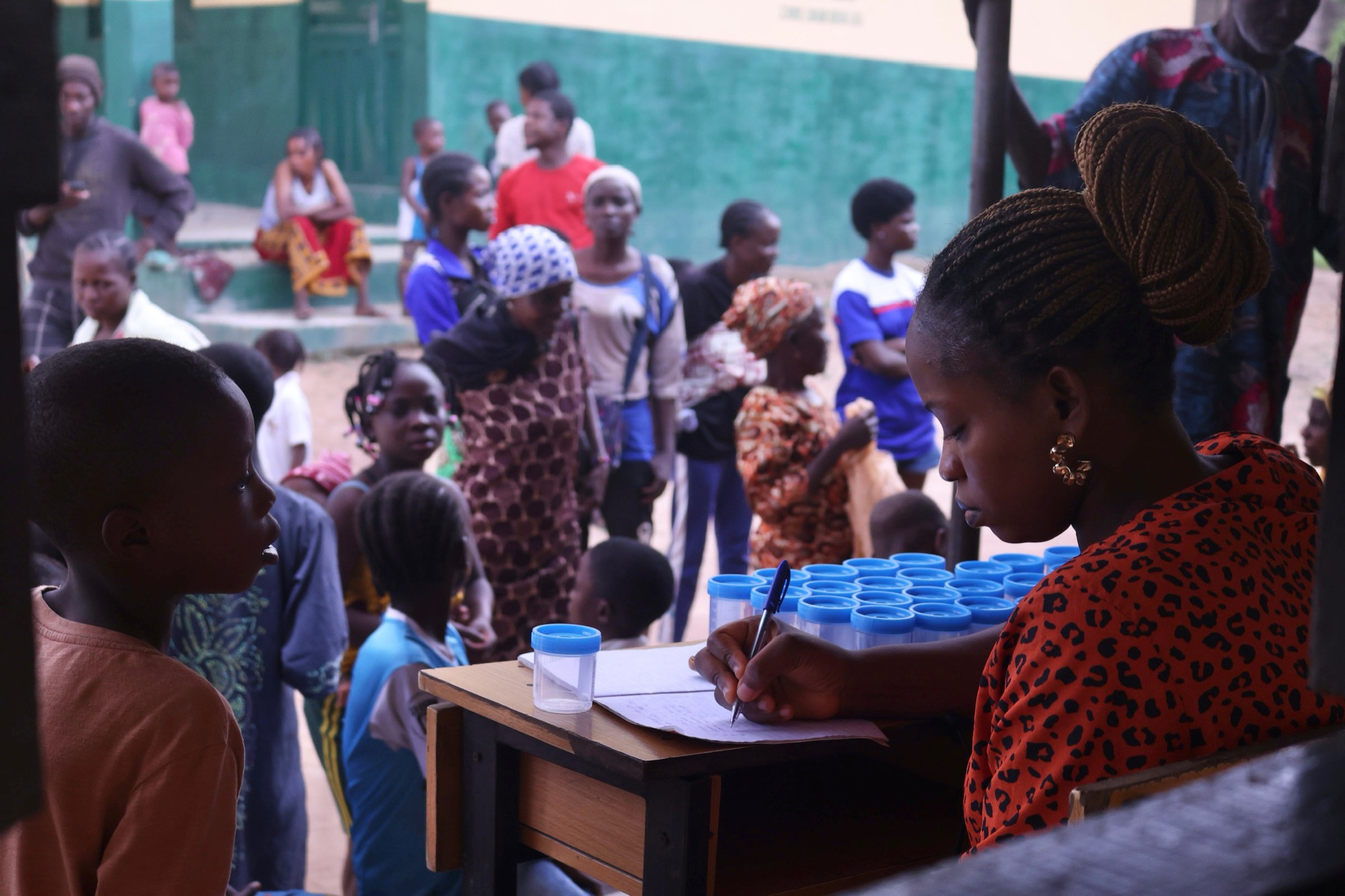 A health worker records participant details at a table of sample cups while a crowd of children and families waits behind her