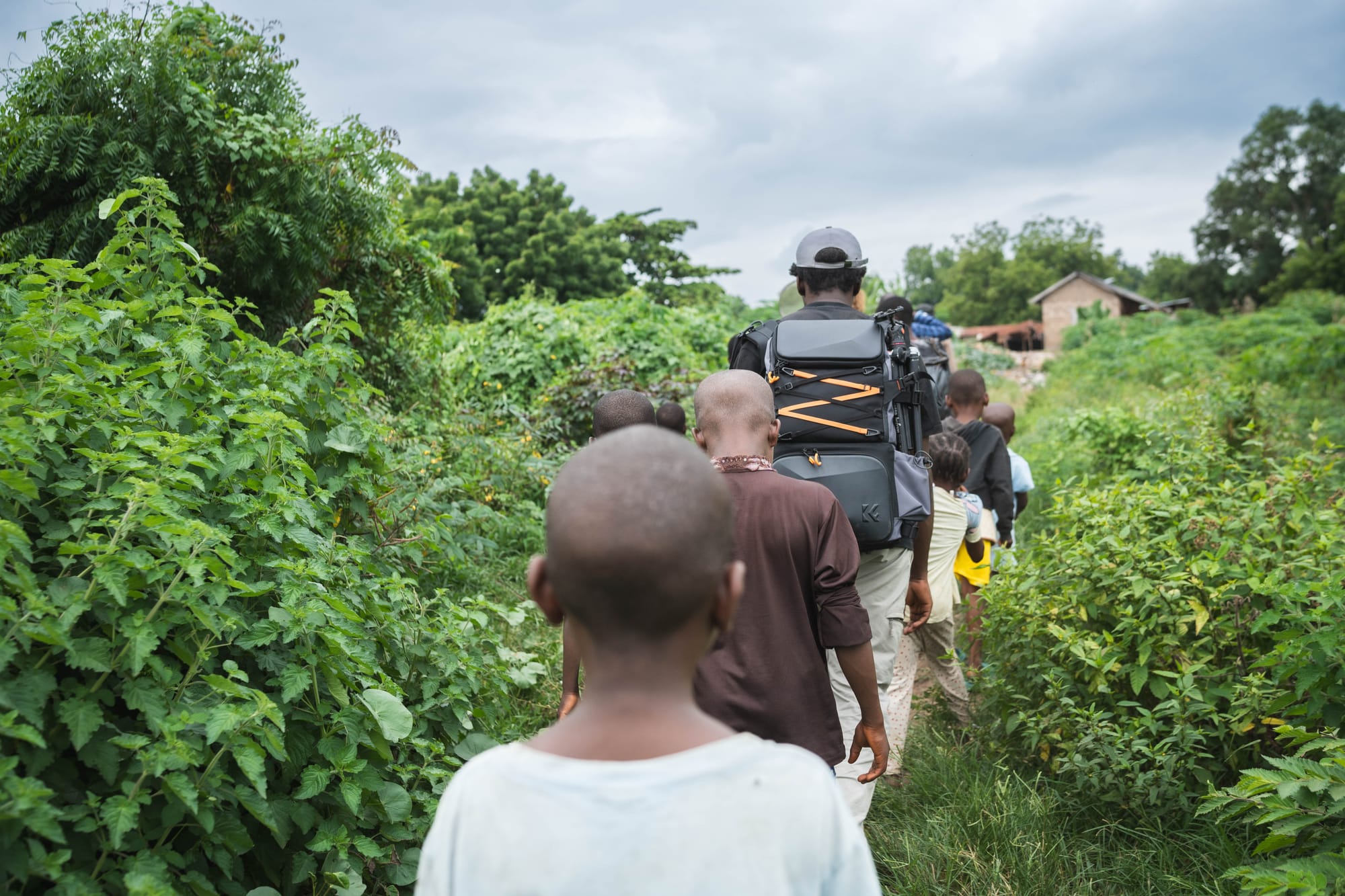 Villagers and the photographer walk single file along a narrow path through tall green brush from the river toward the village