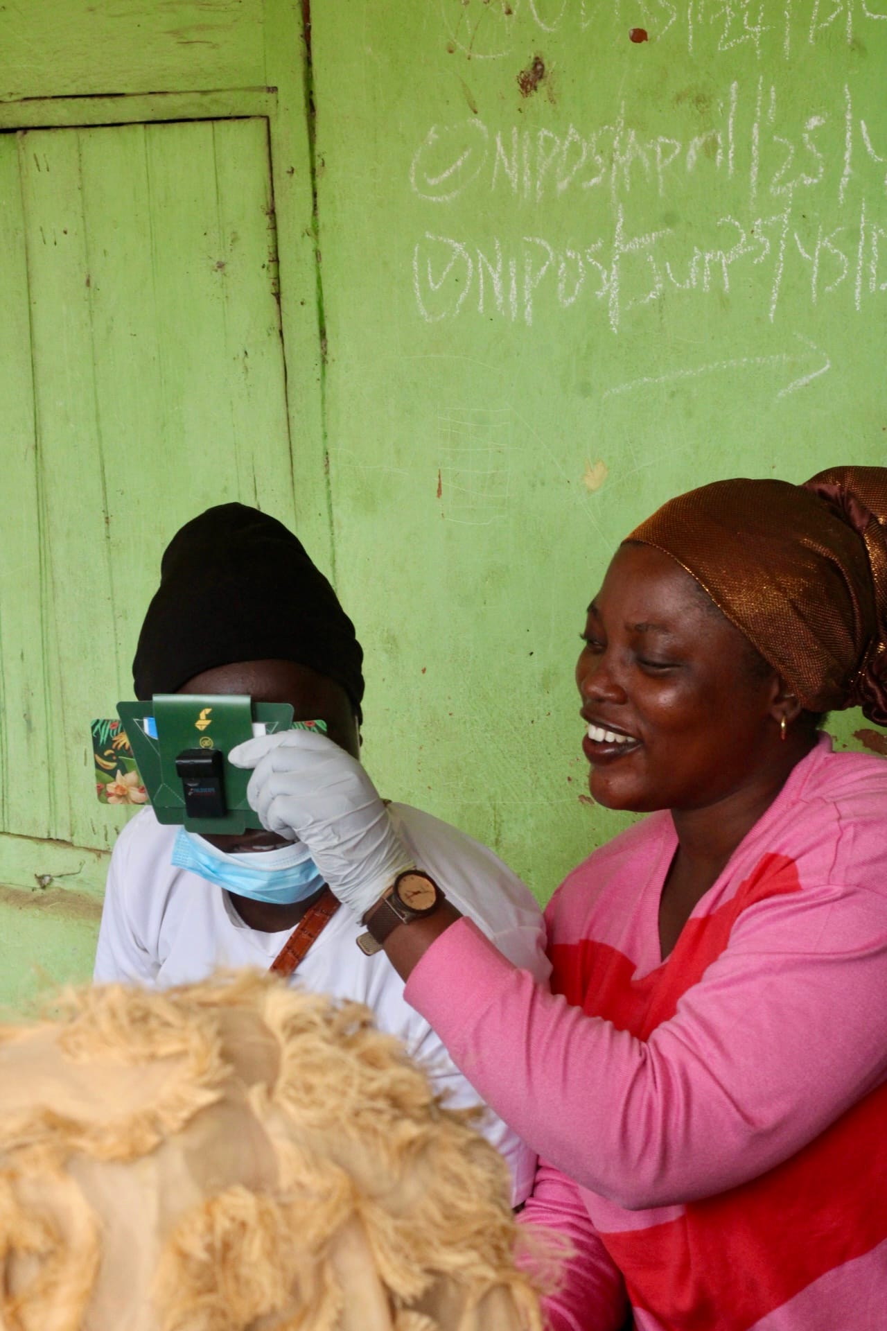 A health worker in a face mask and gloves holds a paper Foldscope up for a smiling woman to look through