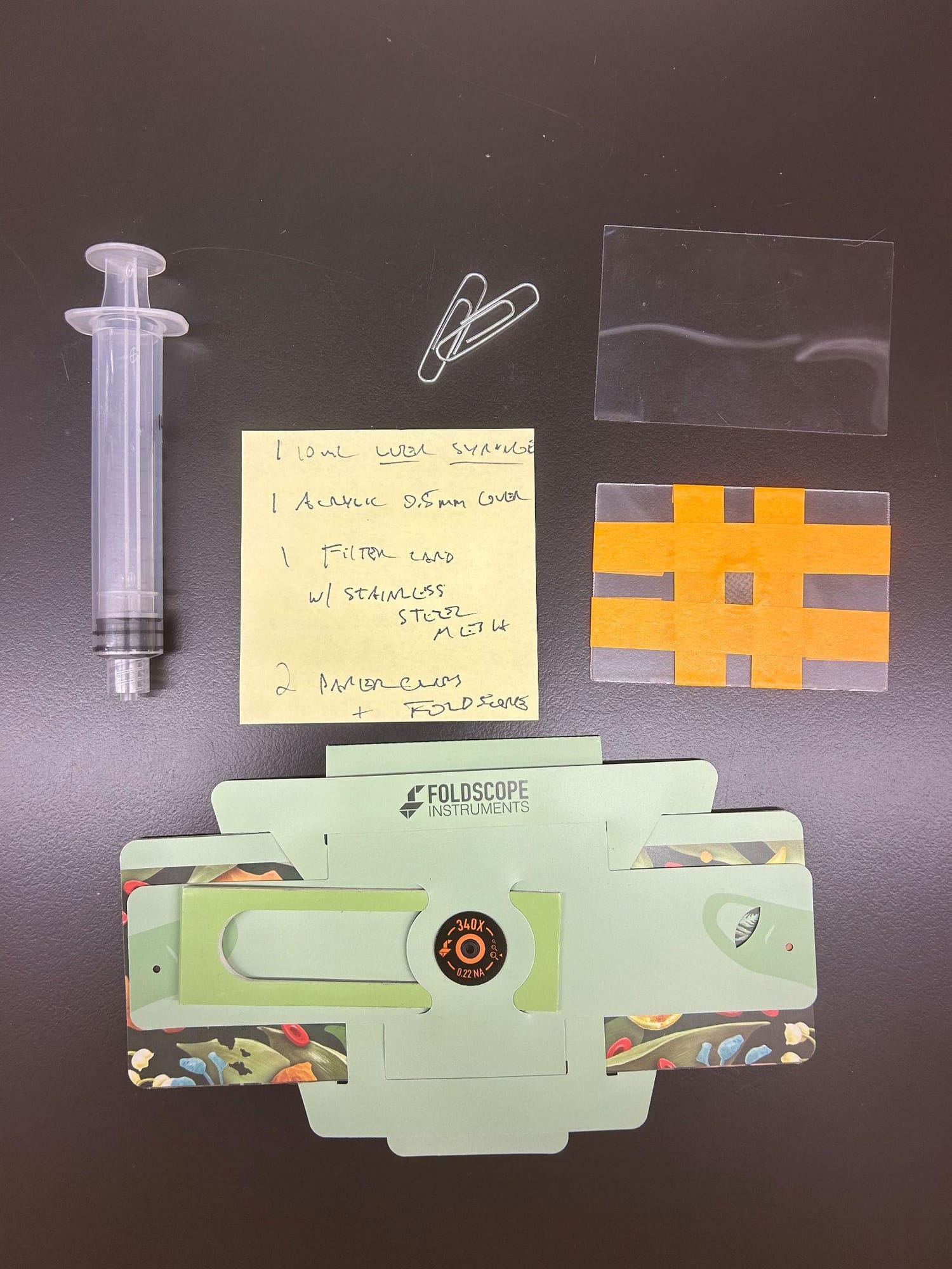 Flat lay of a syringe, paperclips, an acrylic cover, a taped filter card, and a Foldscope, with a sticky note listing the parts