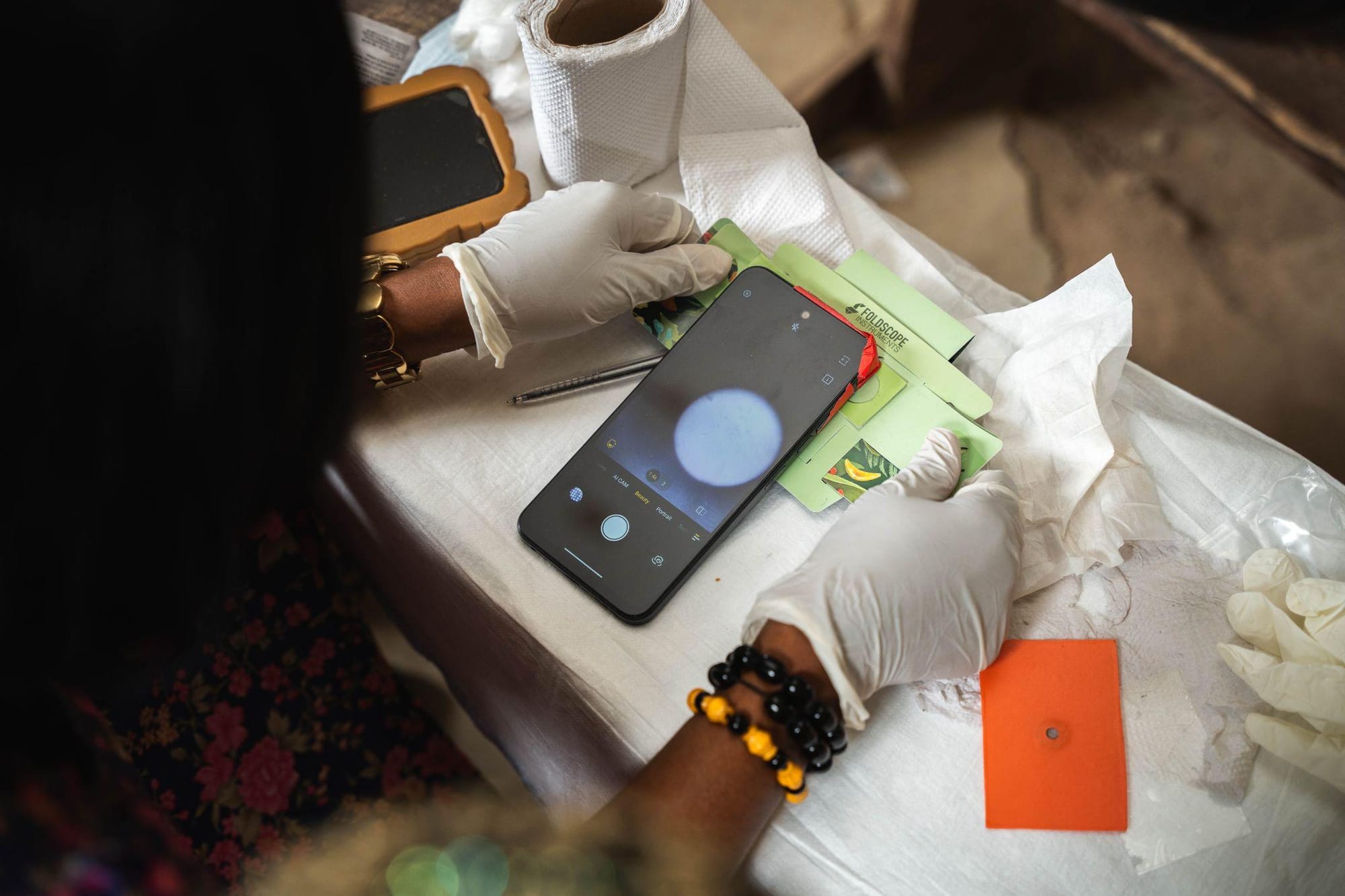 Hands hold orange SchistoFilter cards and a Foldscope over a busy screening table