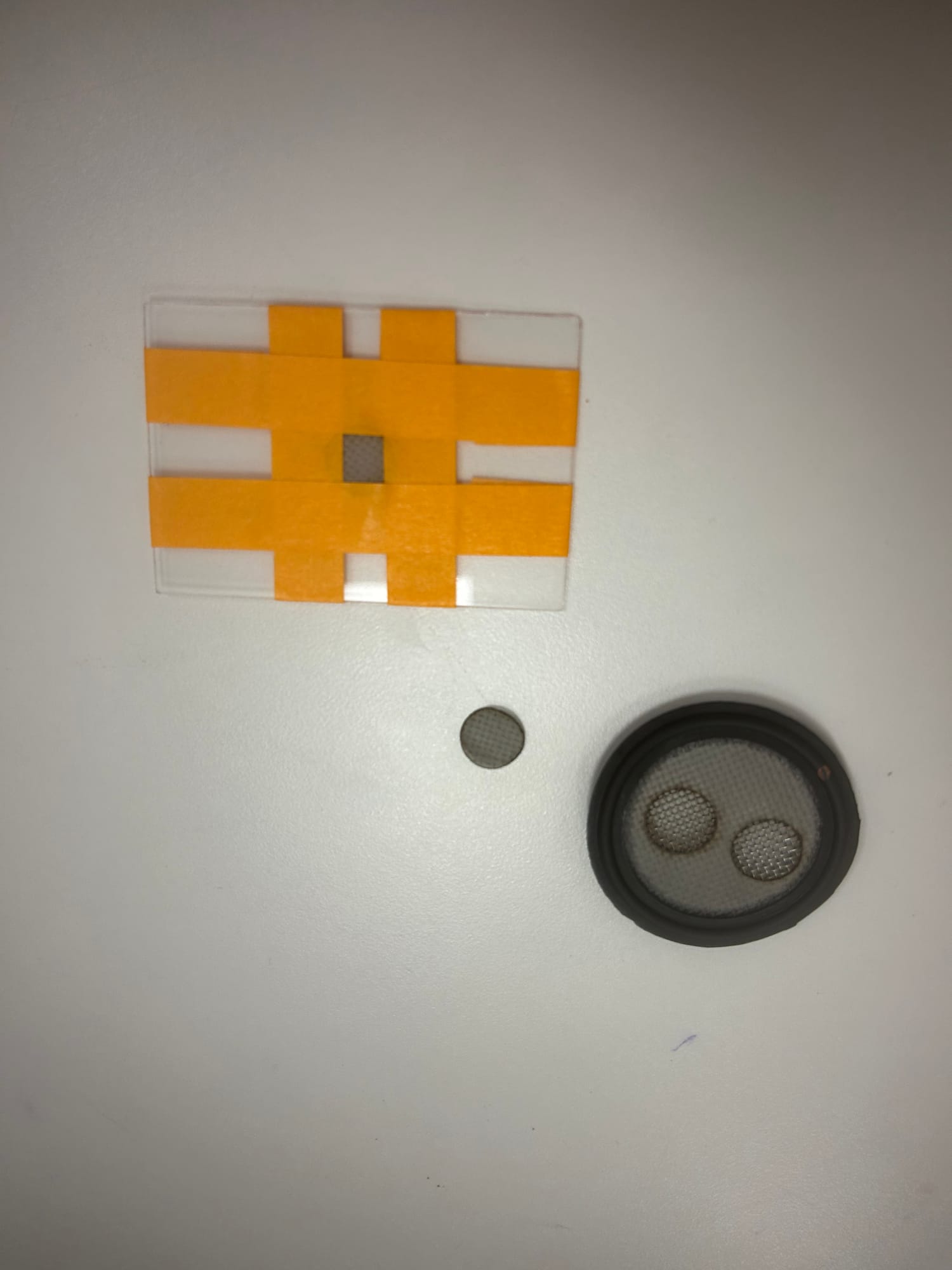 A square of mesh taped into a clear plastic card with orange tape, beside a rubber gasket and a cut disc