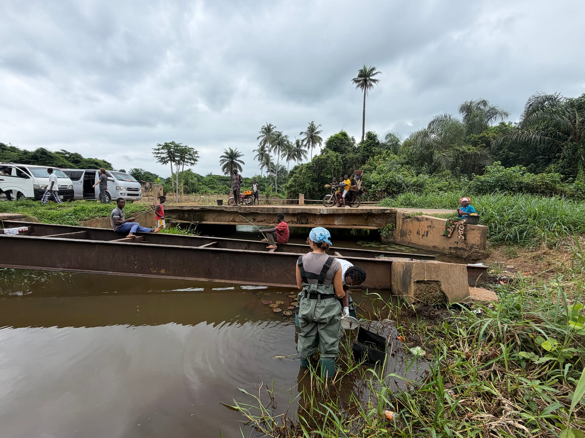 A researcher in chest waders stands in the shallows beside wooden boats and a plank footbridge at a riverside access point