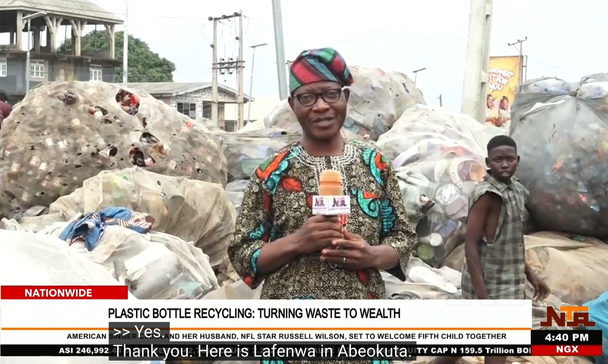 A television news reporter in a patterned cap and dress speaks into a microphone in front of huge sacks of collected plastic bottles