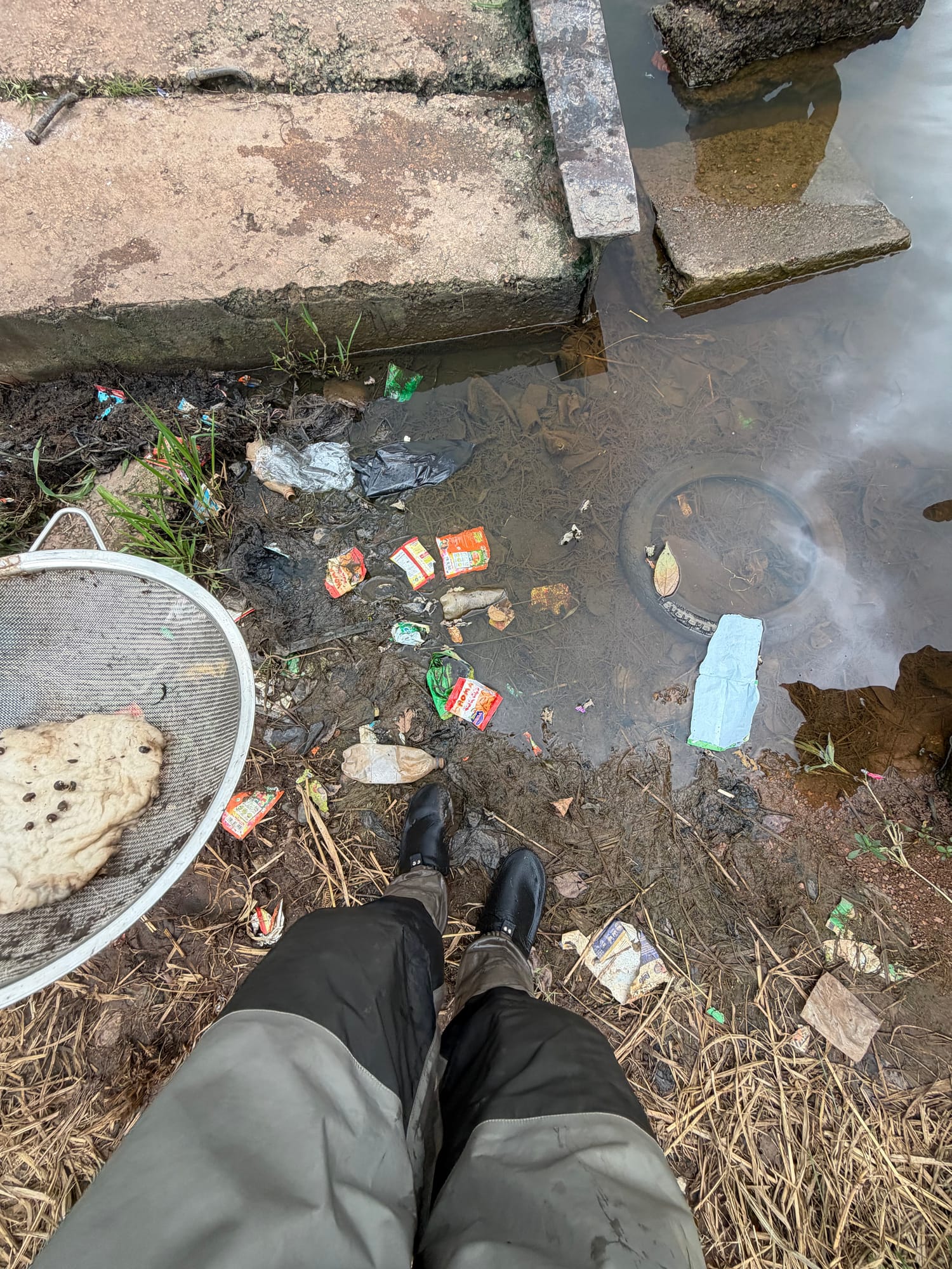 Looking down at waders standing in shallow water littered with plastic, a face mask, and a kitchen sieve