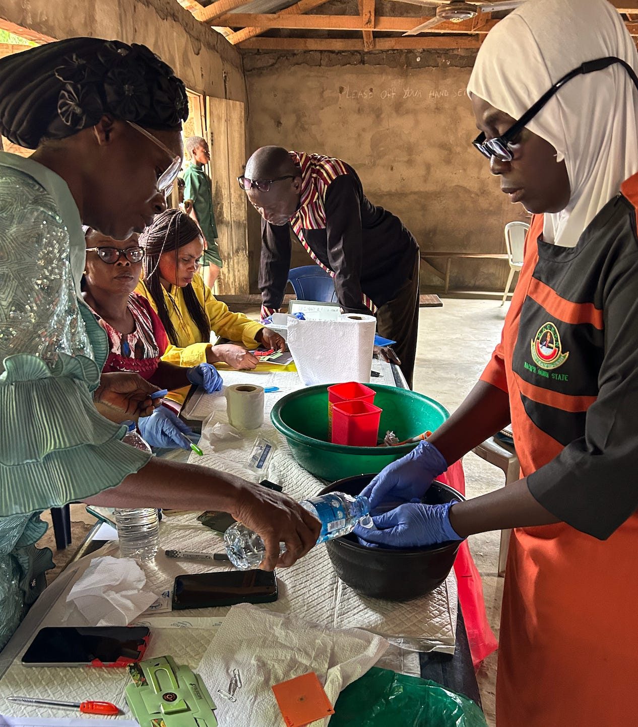 Community health workers in gloves prepare samples around a schoolhouse clinic table with washing bowls and sample cups