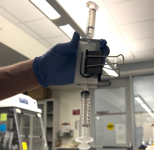 A gloved hand holds the staircase cell upright with syringes above and below and a binder clip
