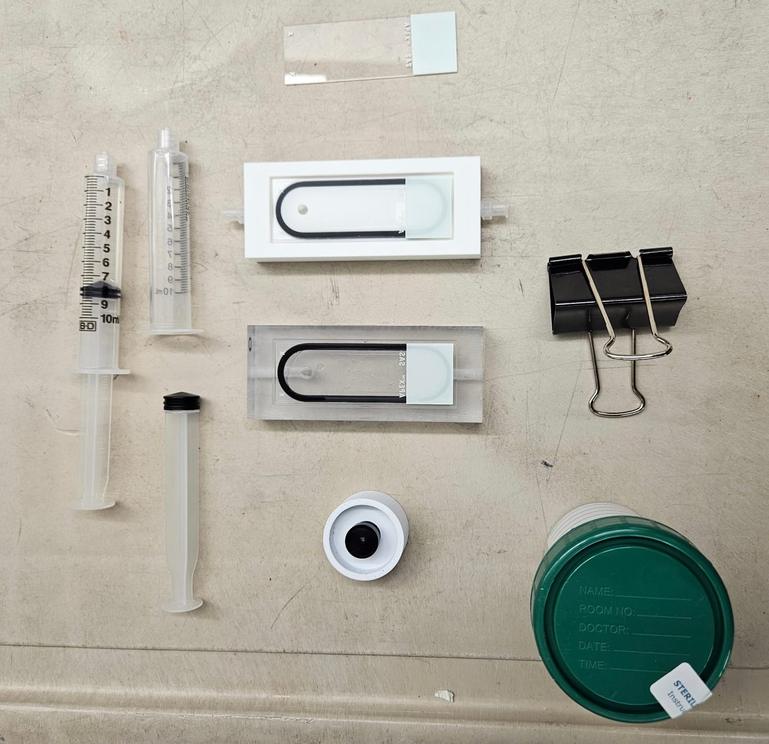 Flat lay of two staircase cell prototypes, syringes, a binder clip, a lens, and a specimen cup